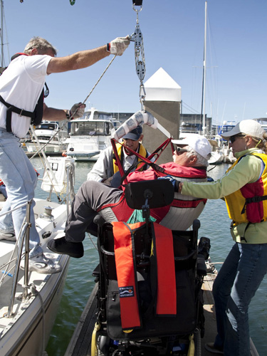 Three volunteers assist in hoisting a sailor from his wheelchair into the keel boat.